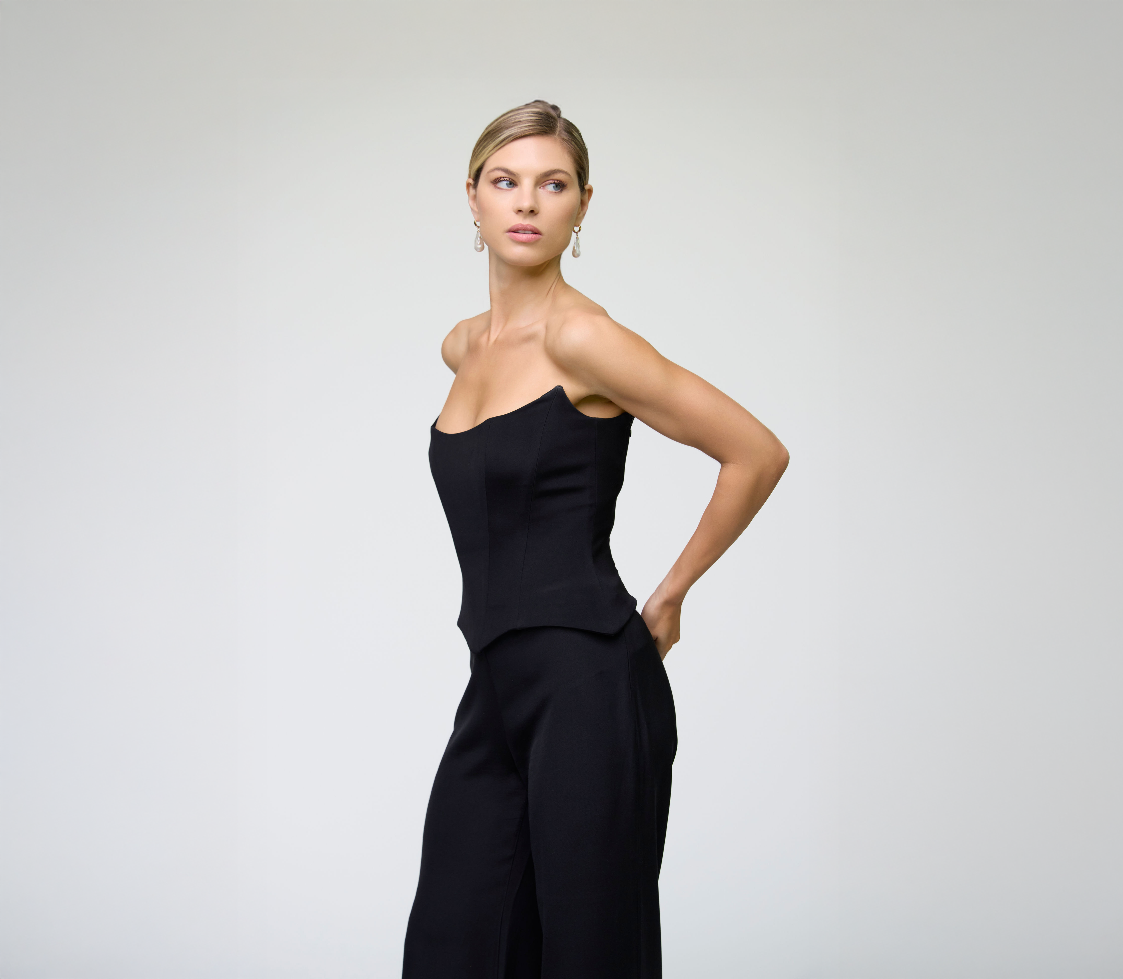 Close-up portrait of a model wearing a black corset top styled with matching trousers against a light grey background.