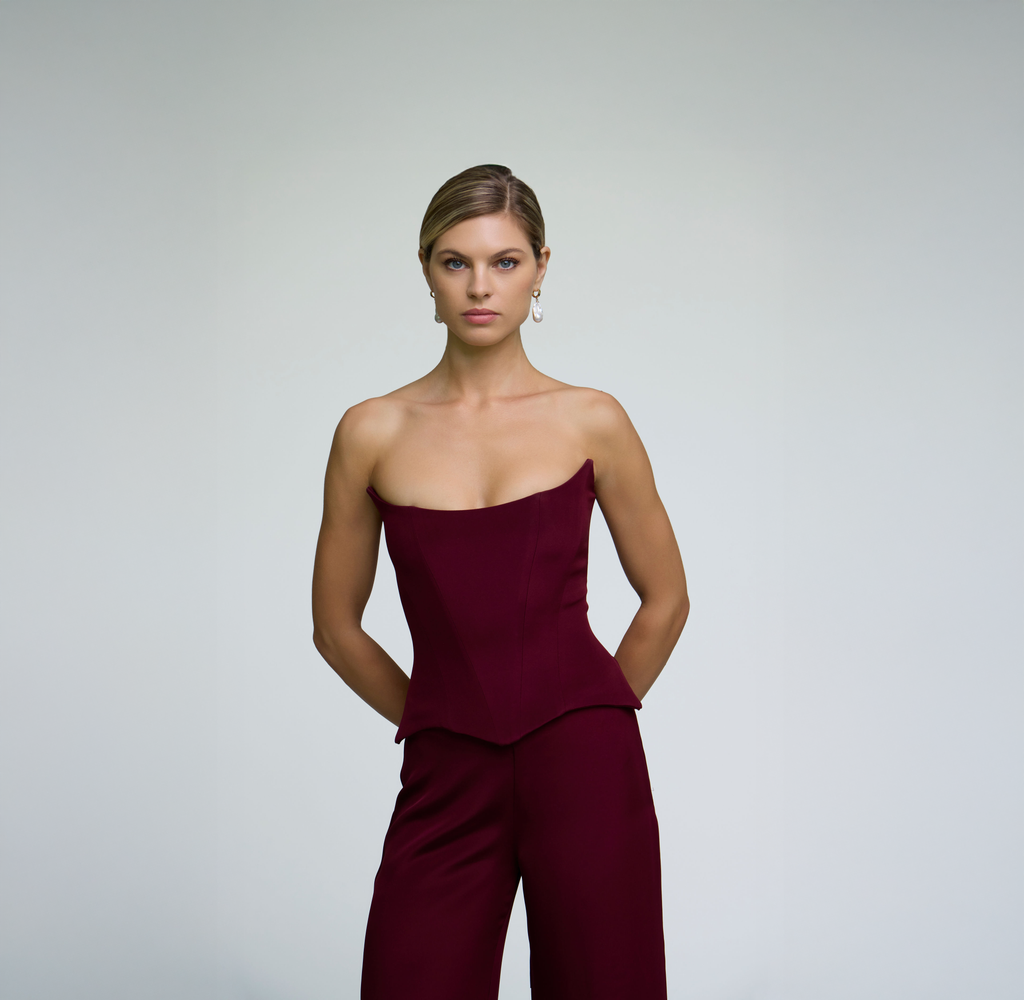 Close-up portrait of a model wearing a burgundy corset top styled with matching trousers against a light grey background.