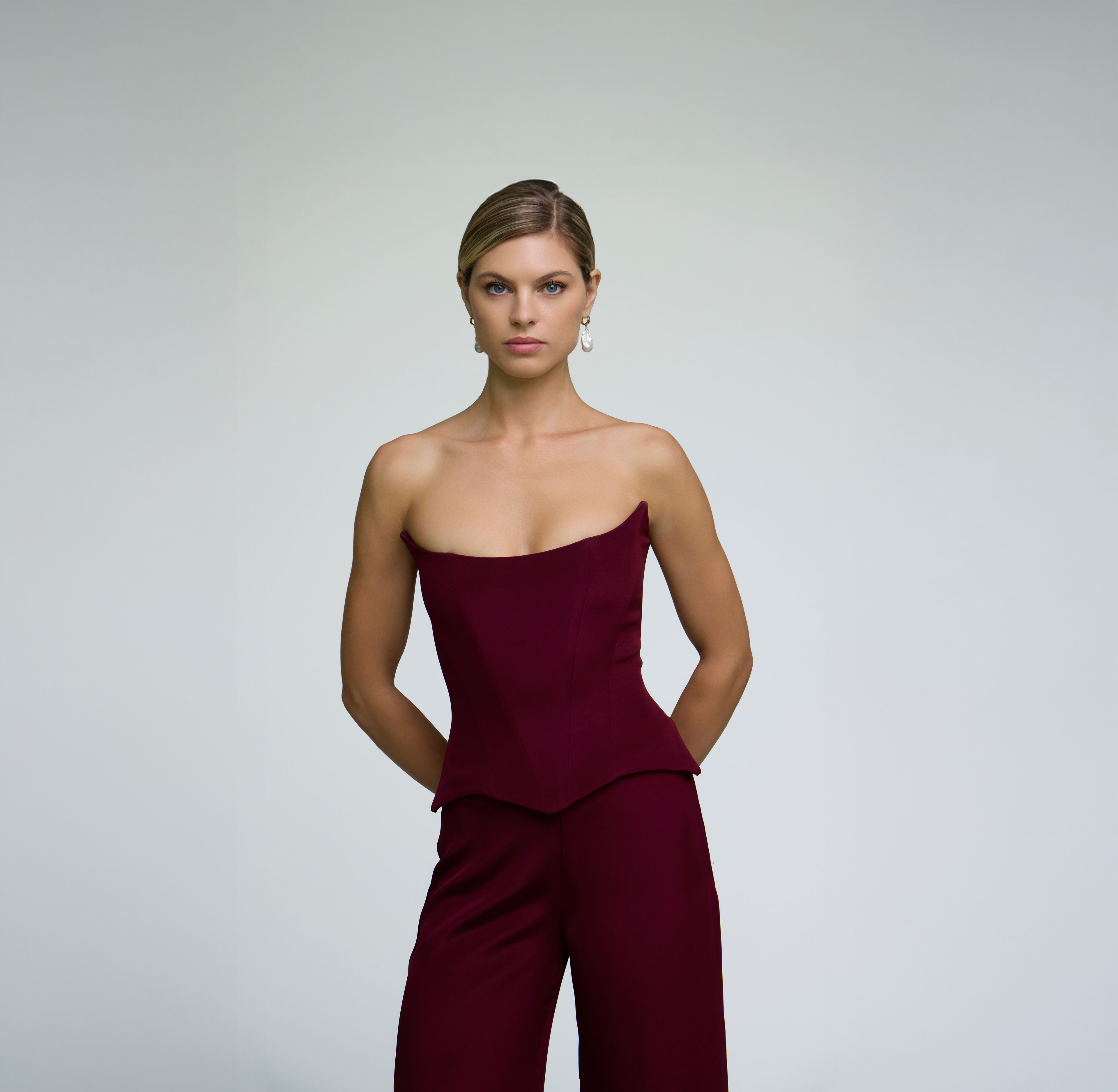 Close-up portrait of a model wearing a burgundy corset top styled with matching trousers against a light grey background.