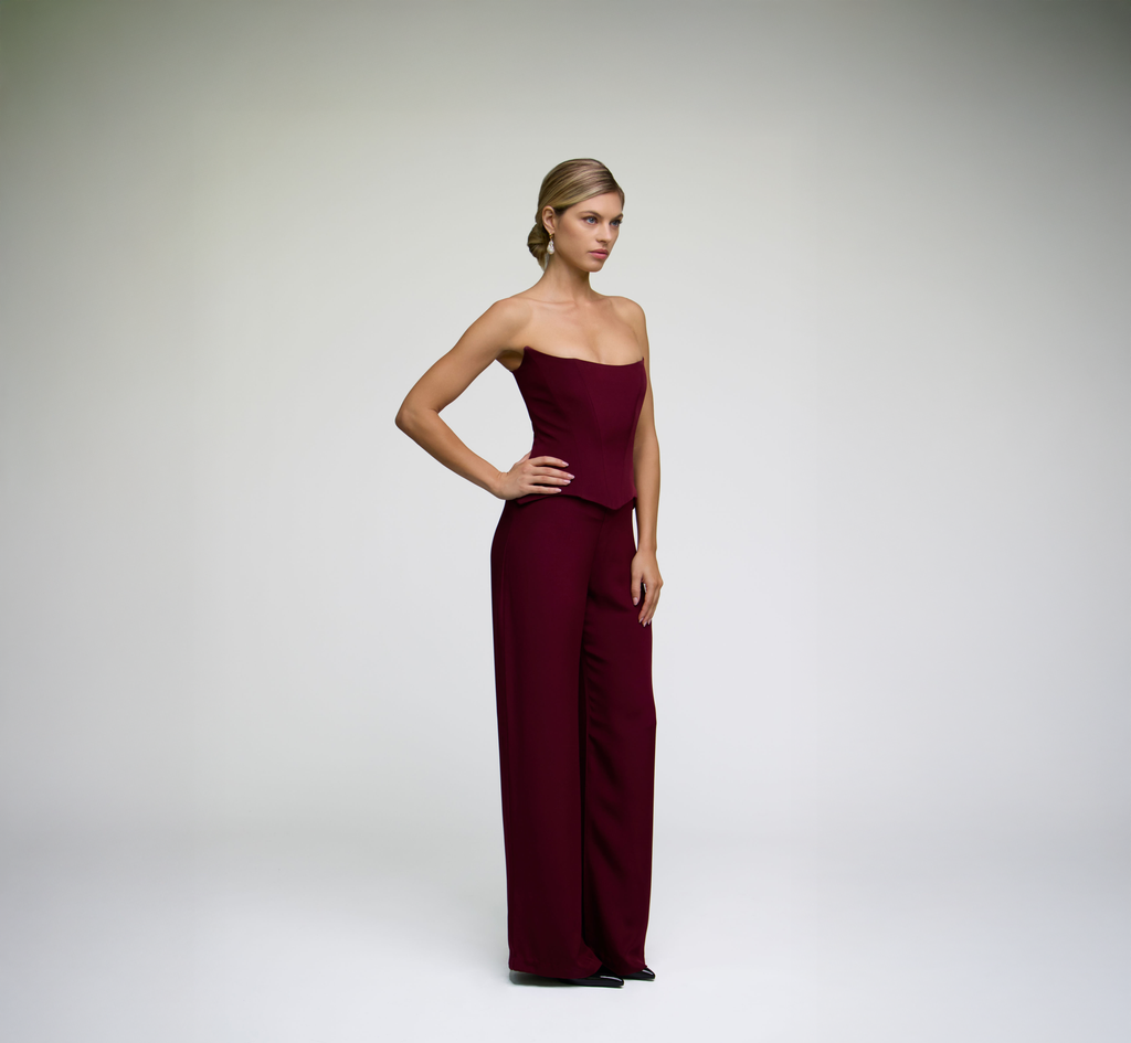 Model posing in a burgundy corset top styled with matching wide-leg trousers at an angle against a light grey background.
