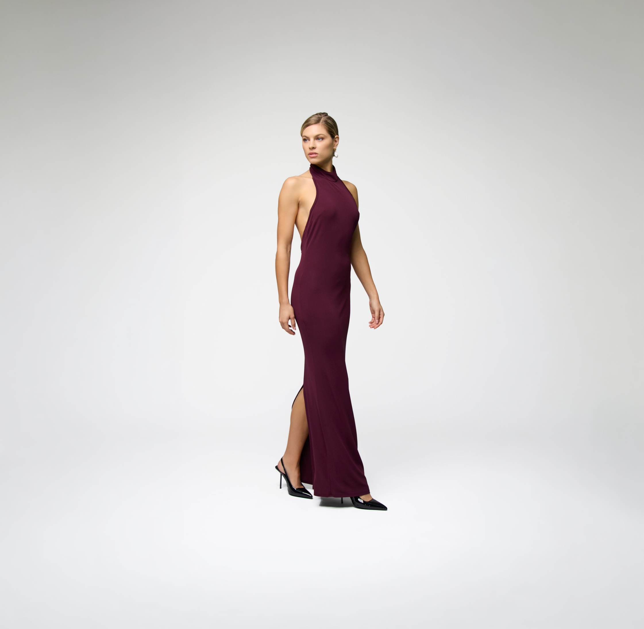 Side view of model wearing ALICHE burgundy halter maxi dress with slit on white background